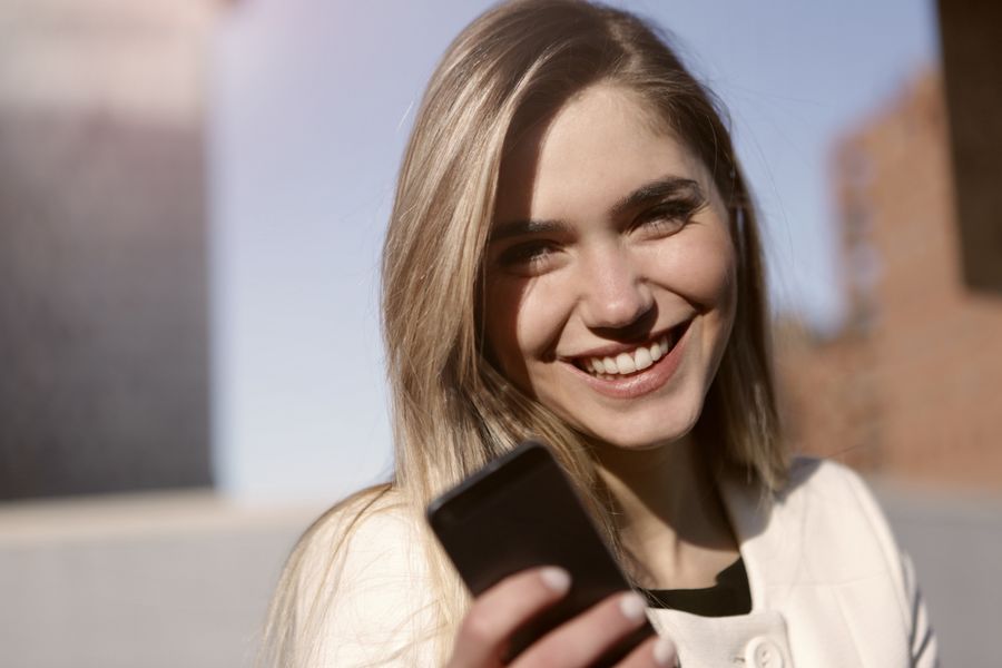 A smiling woman holding a mobile phone outdoors.